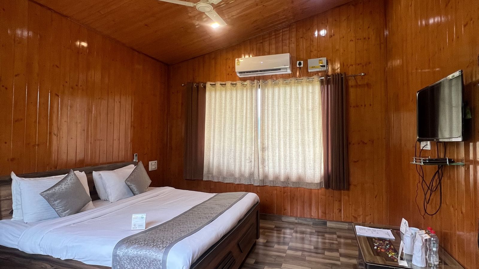 Extended view of the Daksh Elite Cottage, highlighting its wooden interiors, double bed facing a wall-mounted TV, at Daksh The Valley Resort, Saputara.