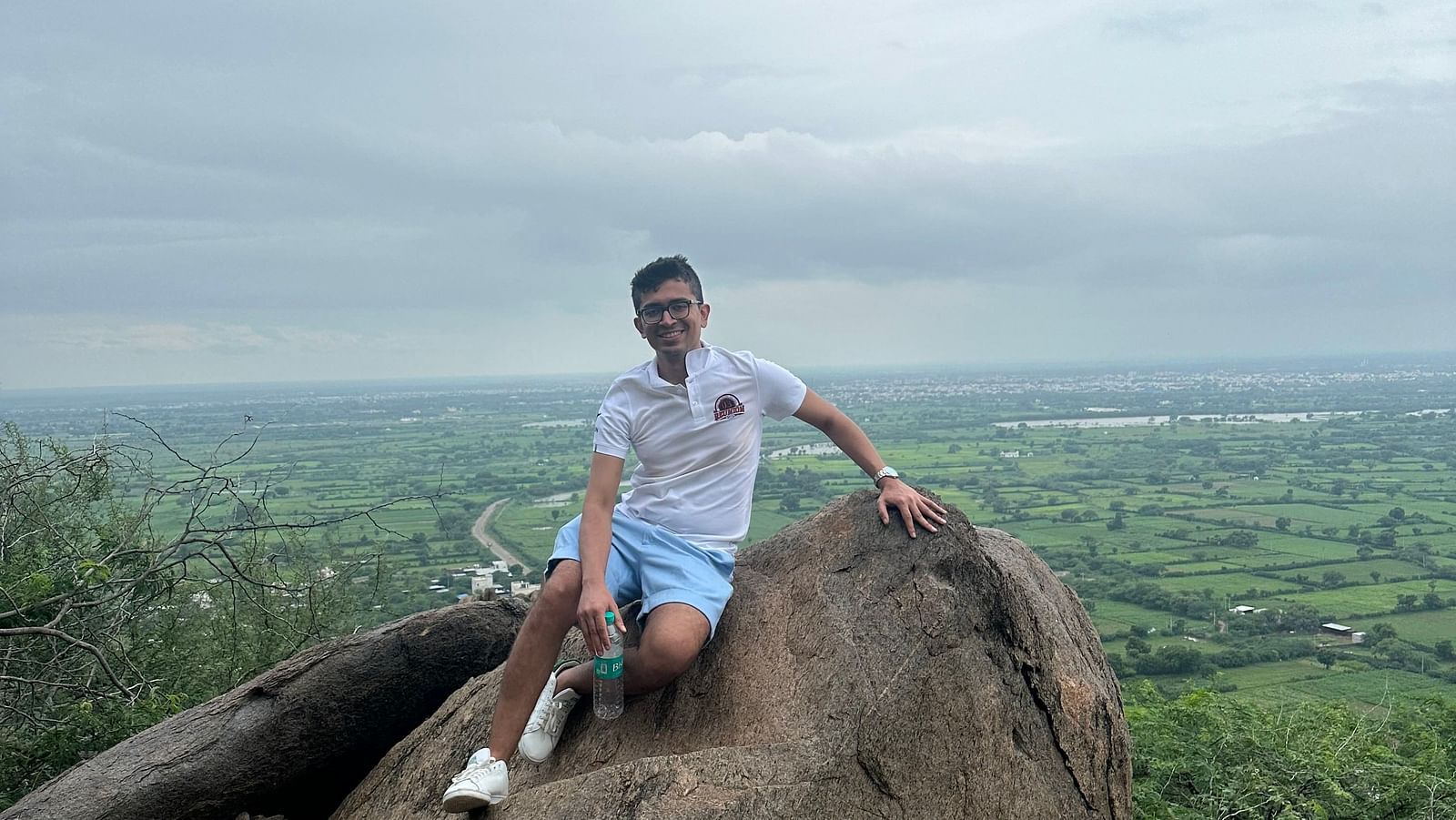 A person sits on a large rock overlooking a landscape with trees and a hazy horizon. 