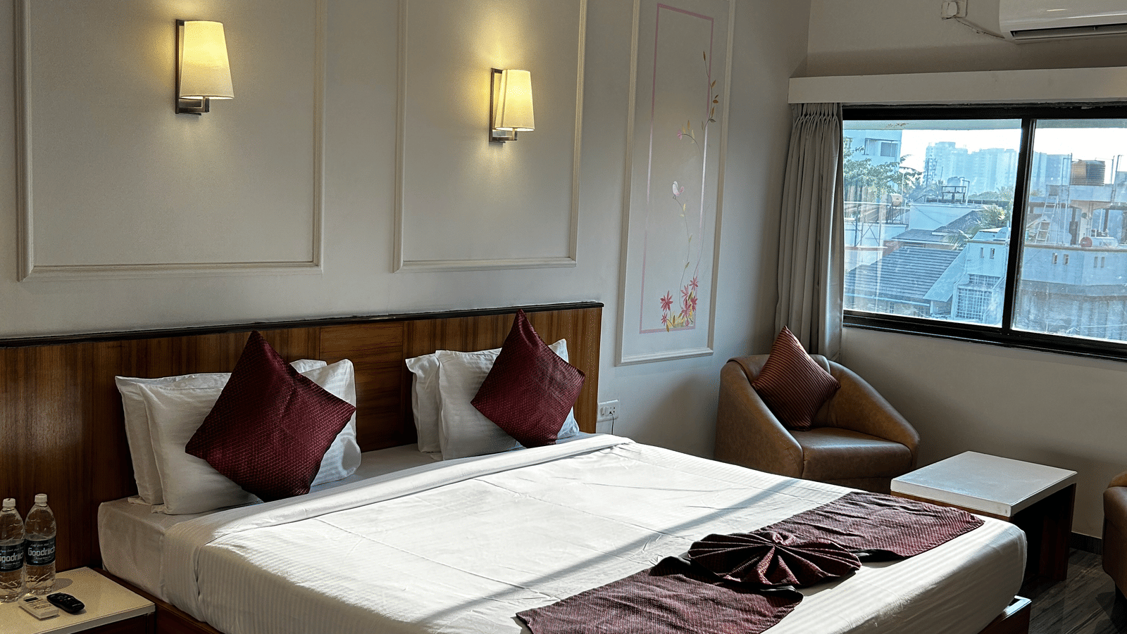  A comfortable guest room at Pravasa Hotel, Kolhapur, featuring a double bed, subtle wall panel moulding, and a wicker armchair.