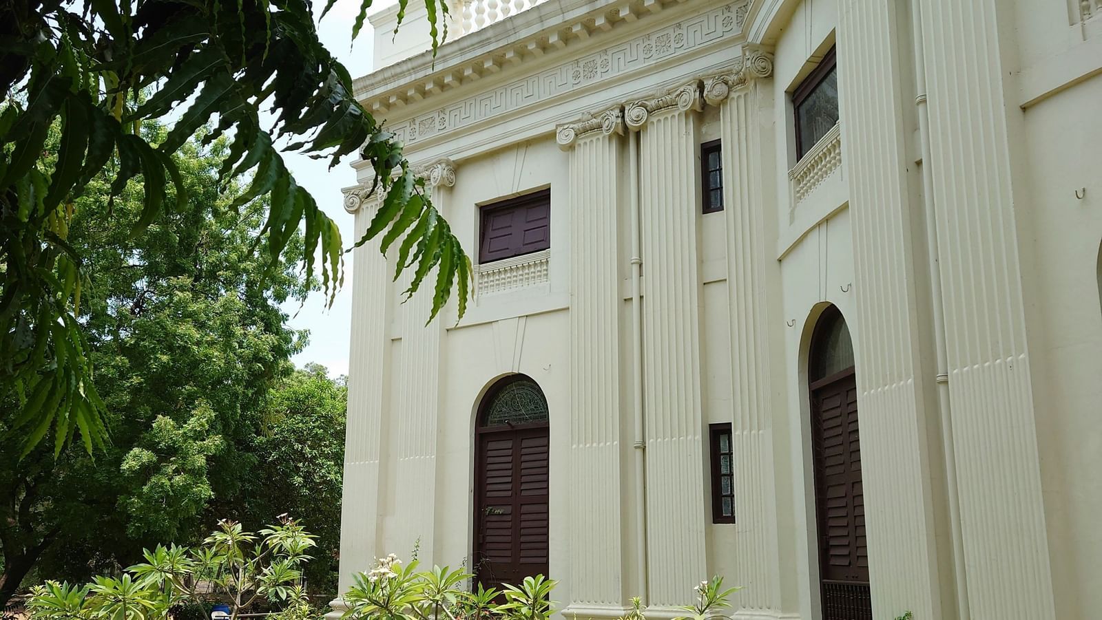 A white heritage-style building with columns and arched windows, surrounded by plants and trees.