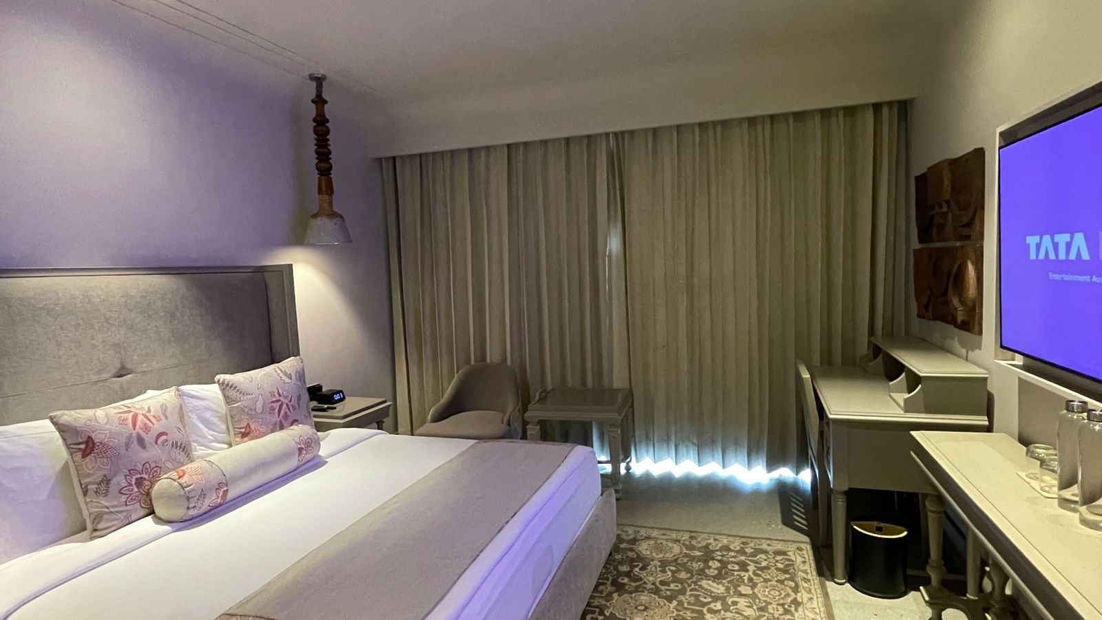 A view of the spacious bedroom at The Evren, Vagator, with a large bed featuring decorative pillows and a bed runner. The room has a patterned rug, a desk, a mounted television, and a window with curtains.