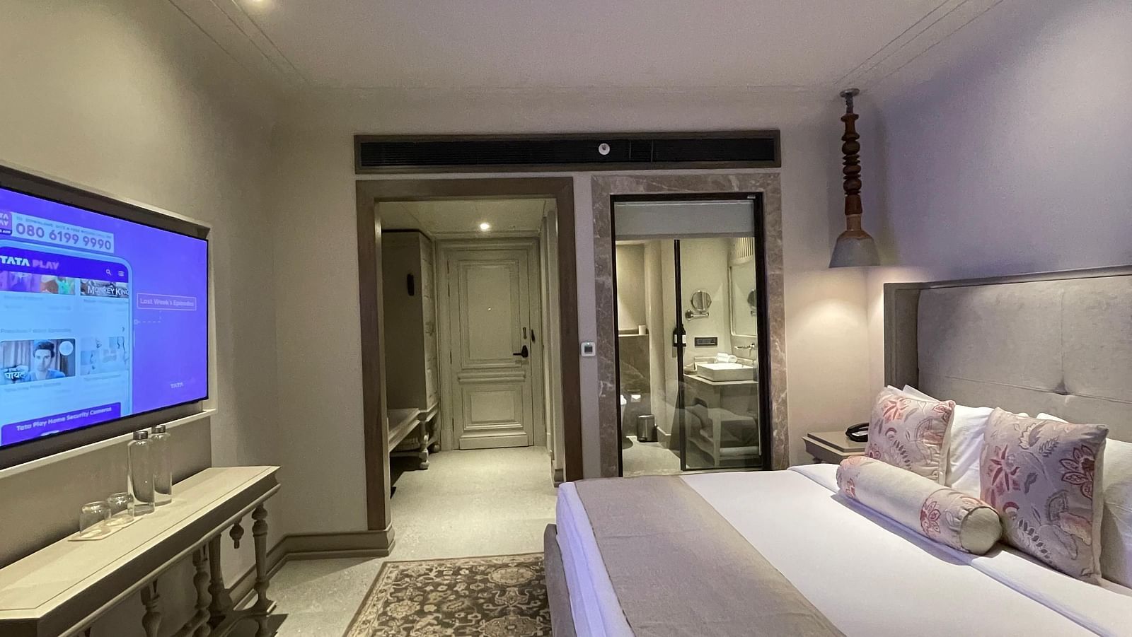 A wide-angle view of the bedroom at The Evren, Vagator, showing the bed, television, and the open entryway leading to the bathroom. A patterned rug covers a portion of the floor.