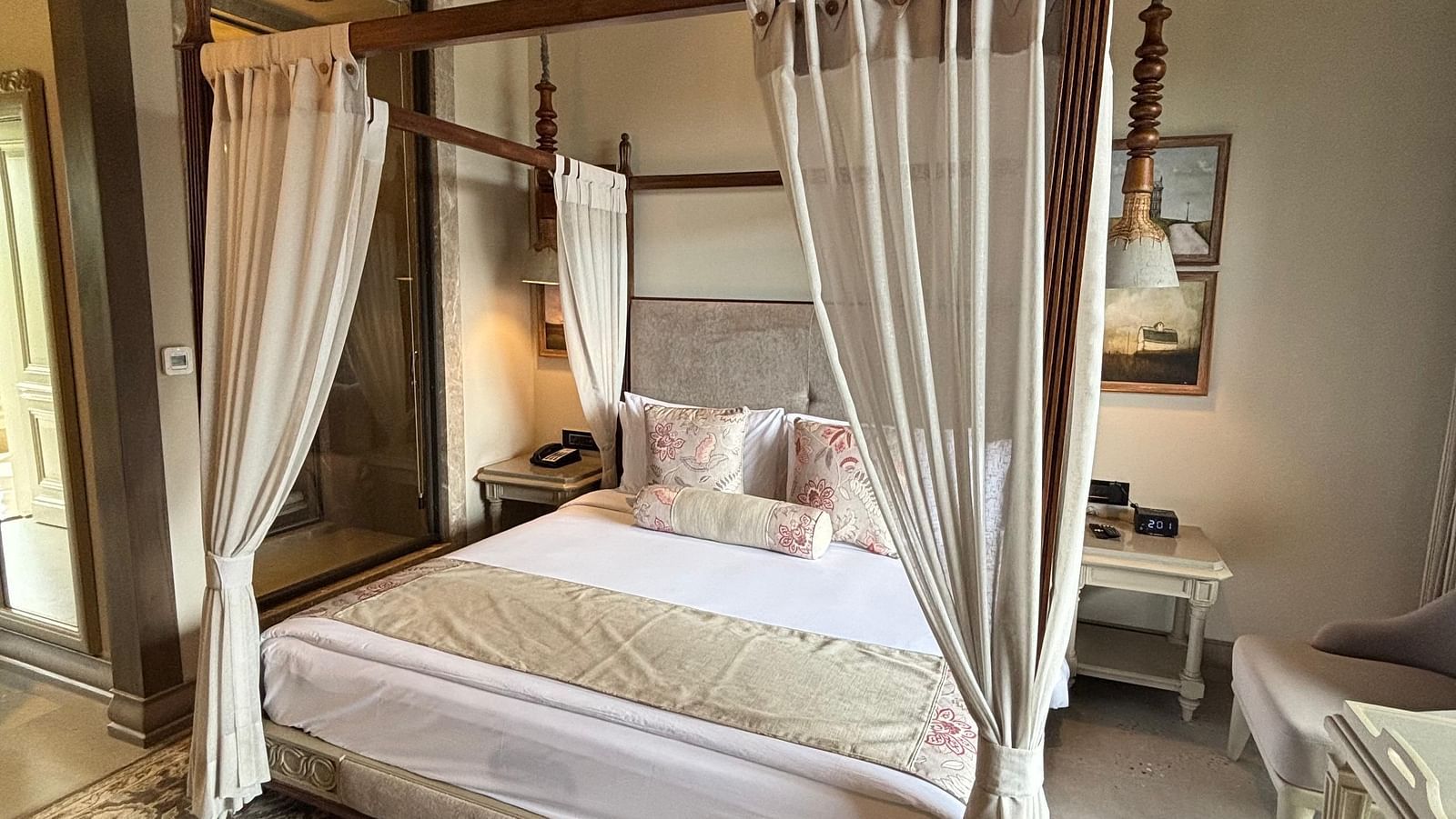 A wide shot of a Premium King Room at The Evren, Vagator, featuring a four-poster bed with elegant curtains and a patterned rug. The room has a high, sloped wooden ceiling and an open doorway leading to the bathroom.