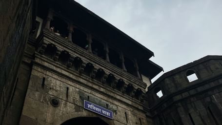 A low-angle shot of the corner of a grand, traditional Indian building, with a balcony and an arched entryway below, set against a cloudy sky.