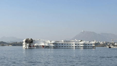 A wide scenic view of the calm waters of Lake Pichola in Udaipur, with the Aravalli Hills visible in the misty distance.