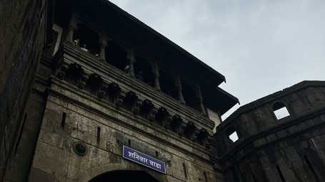 A low-angle shot of the corner of a grand, traditional Indian building, with a balcony and an arched entryway below, set against a cloudy sky.