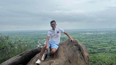 A person sits on a large rock overlooking a landscape with trees and a hazy horizon. 