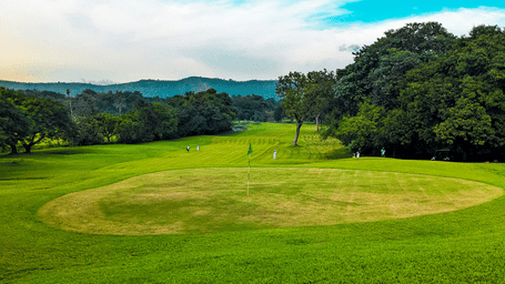 Expansive golf course with rolling fairways, vibrant greenery and distant hills beneath a bright blue sky with scattered white clouds.