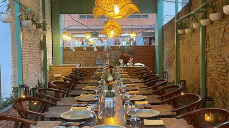 A long dining table set with place settings and wooden chairs beneath warm woven pendant lights in a narrow semi-outdoor dining corridor at  The Nanee, Bhaktapur