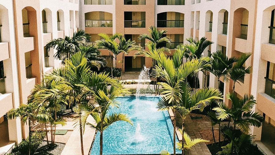 A view of a resort courtyard with a central pool, palm trees lining the walkway, and cream-coloured buildings framing the tropical setting.