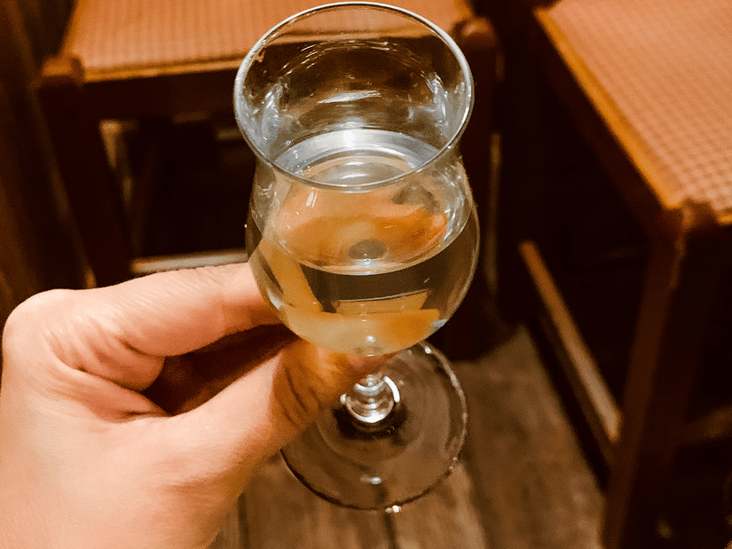 Hand holding a small glass of clear spirit on a wooden table inside a warmly lit rustic bar setting