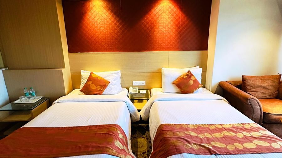  A modern hotel room at Amer Greens featuring two twin beds with white linen and burnt orange runners, flanking a nightstand.