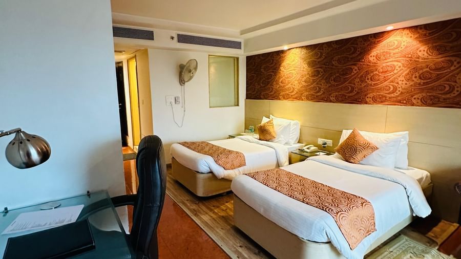 A bright indoor view of a hotel room at Amer Greens featuring two beds, a patterned accent wall, and a desk area with an office chair.