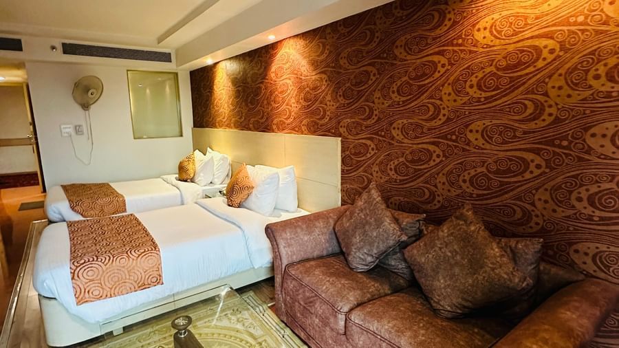  An indoor view of a hotel room with two beds and a sofa, at Amer Greens, featuring a patterned accent wall and overhead lighting.
