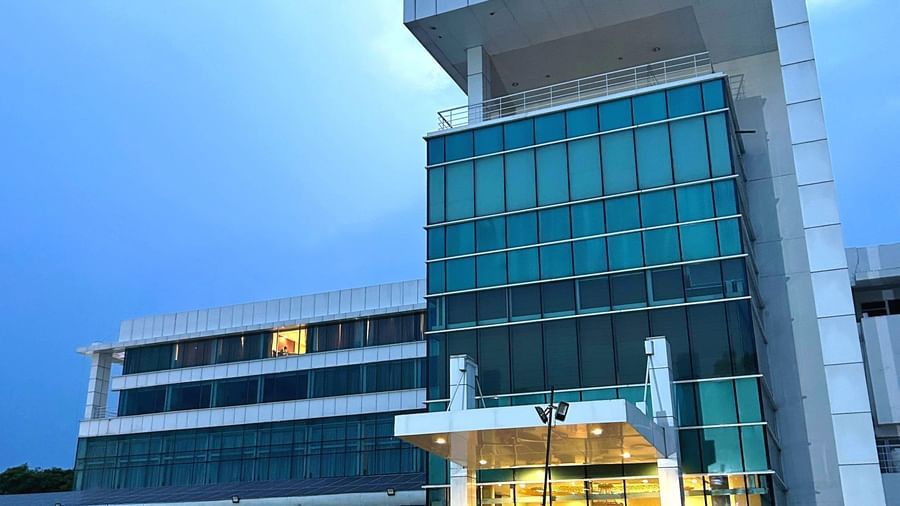  An angled exterior view of the modern, glass-fronted Amer Greens hotel building at dusk, featuring a bright entrance and manicured hedges.