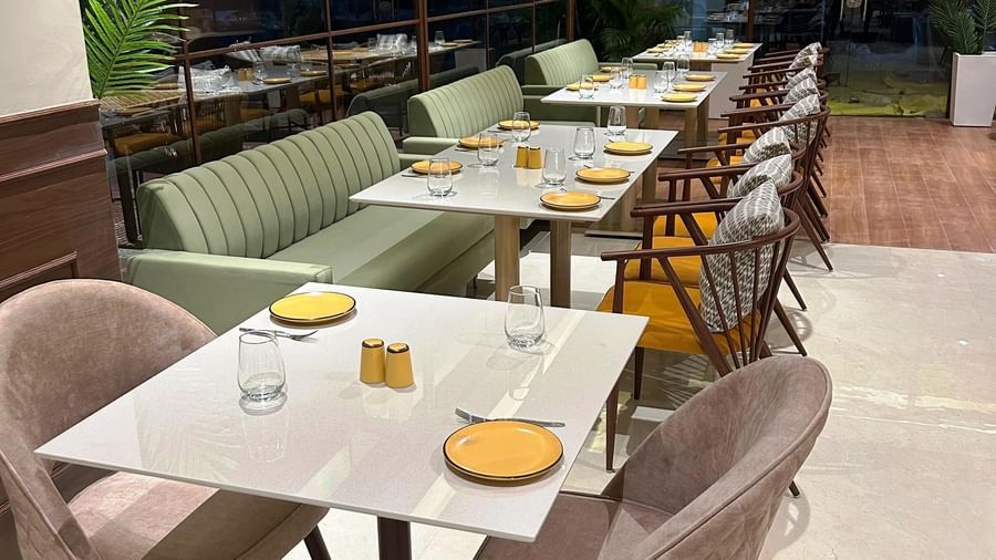 An indoor dining area at Amer Greens featuring rows of tables and chairs, with a long built-in sofa along a wall of tall windows looking out into the night.