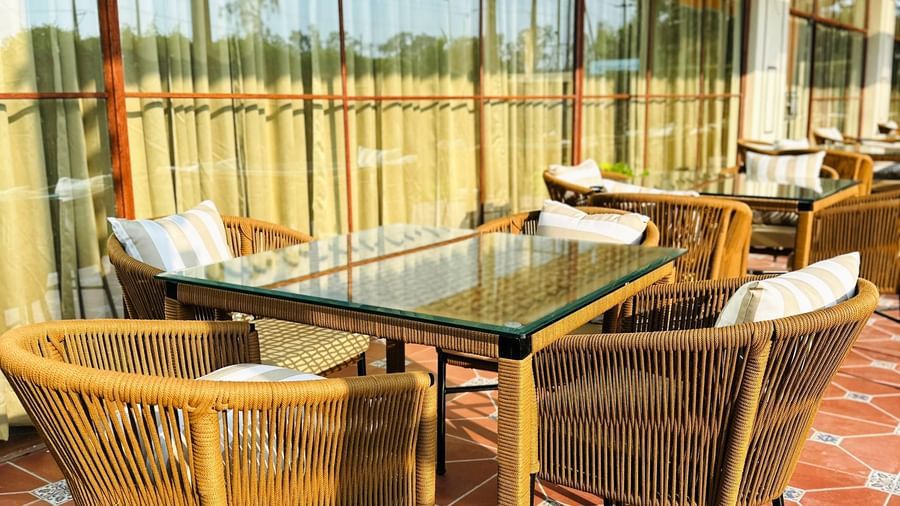  An outdoor seating area at Amer Greens featuring several glass-top square tables and woven chairs with cushions, set on a patterned tile floor next to tall windows.