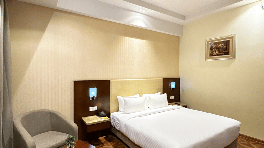 Wide view of Deluxe Bedroom at Golden Tulip Jaipur, Kukas, featuring a king size bed, an upholstered armchair, and wooden flooring.