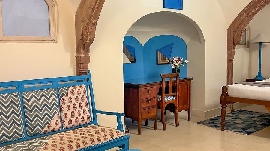 A hotel suite with a seating area featuring a blue sofa and coffee table, arched architectural details, and a view into a bedroom with a bed and a desk in another alcove at Tijara Fort-Palace - 19th Century, Alwar.