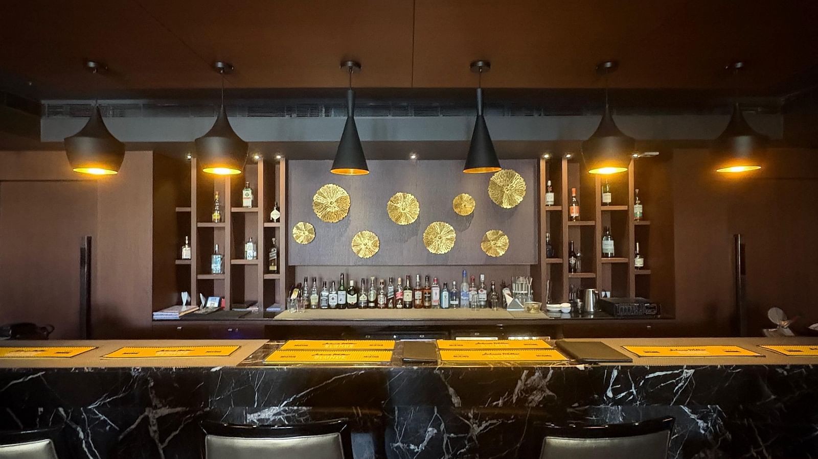 A dimly lit bar with a dark, textured wall and a row of bottles illuminated from behind, highlighting the bar's moody atmosphere.