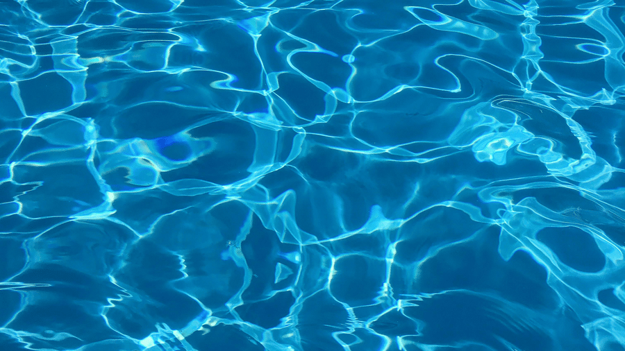 A close-up vertical shot of bright, clear, azure blue water in a swimming pool, with shimmering light reflections on the surface.
