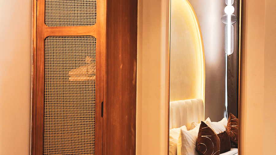 A wooden wardrobe with mesh panels stands next to a tall, thin mirror reflecting a bed with brown cushions at Avanya Resorts N Retreat.