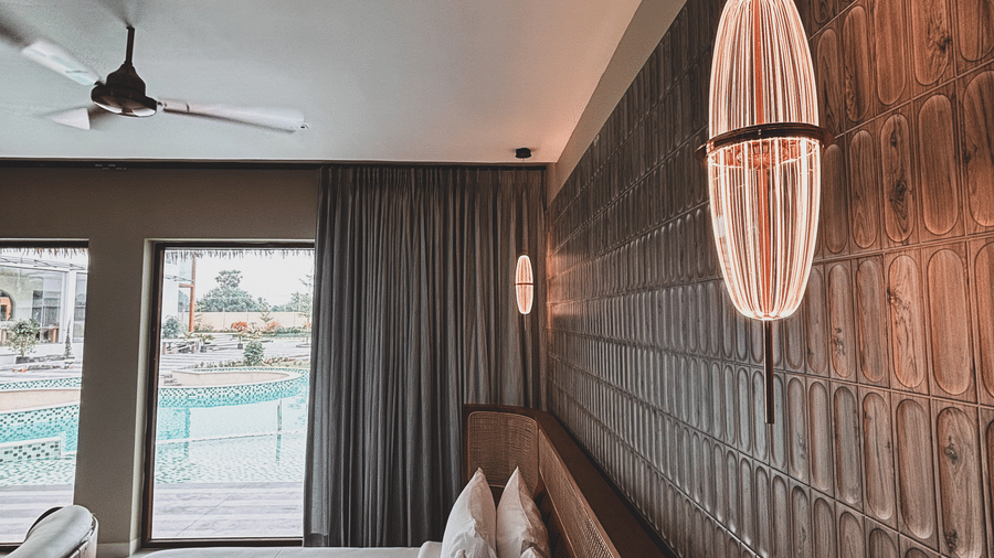 A vertical oval light fixture is mounted on a textured brown wall next to a bed with 4 white pillows at Avanya Resorts N Retreat.