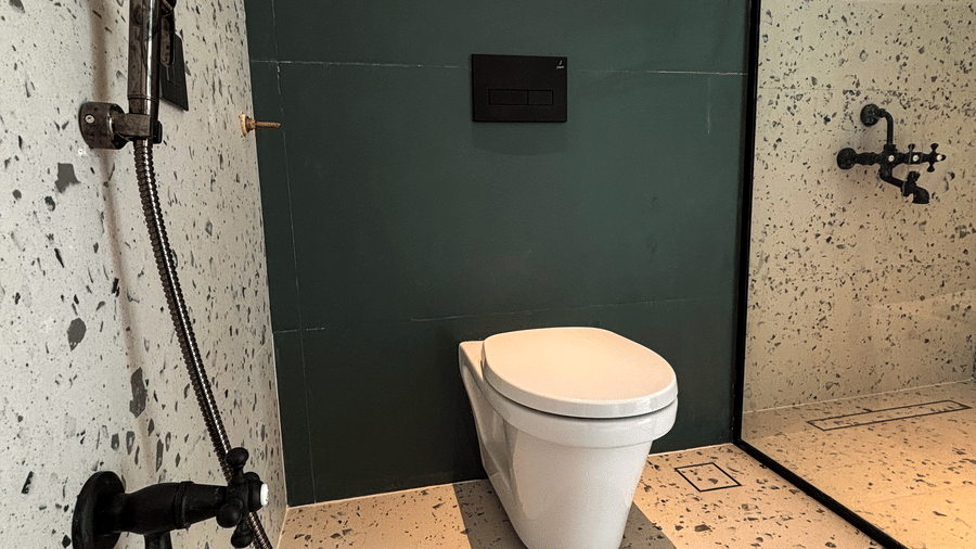 A white wall-mounted toilet is set against a dark green panel and surrounded by speckled terrazzo tiles at Avanya Resorts N Retreat.