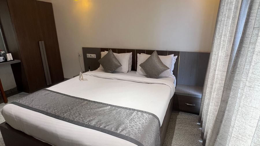 Corner view of Daksh Comfort room at Daksh The Valley Resort, Saputara, featuring a cosy room with a plush king size bed, a wardrobe, warm overhead lighting, and long closed curtains on one side of the room.