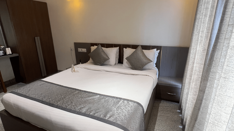 Corner view of Deluxe Room with Road View at Daksh The Valley Resort, Saputara, featuring a cosy room with a plush king size bed, a wardrobe, warm overhead lighting, and long closed curtains on one side of the room.