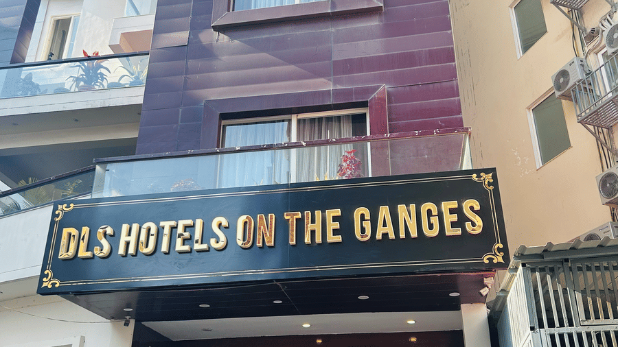 Vertical exterior view of DLS Hotels on the Ganges, highlighting the building’s height, the entrance below, and the large yellow hotel sign, framed by adjacent structures, showing the prominent and accessible hotel entrance