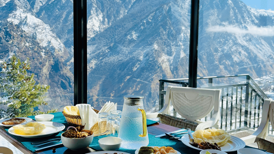  Breakfast spread on a table inside a glass dining area with snowy mountain views at Faraway Cottages, Auli.
