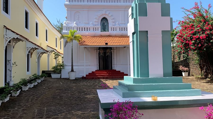  A vibrant outdoor scene at Fort Tiracol, Goa, showing a white building with a red and white structure, possibly a statue or a decorative element, and a potted plant in the foreground.