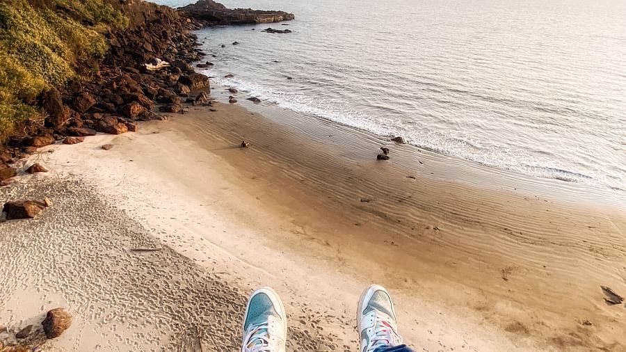 A first-person perspective shot looking down at two legs in blue jeans and white sneakers, with a sandy beach and the ocean visible in the background, suggesting someone is sitting or lying down on a elevated position overlooking the water.