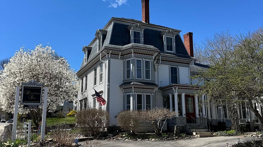 Hartstone Inn on a sunny day, showcasing its grand Victorian architecture, American flag on top, and blooming trees lining the driveway.