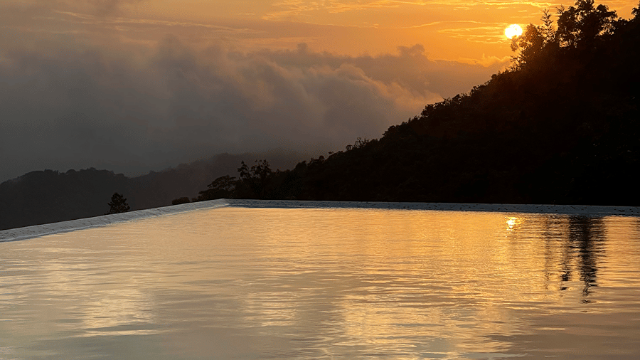 The sun setting behind a hill, reflecting off the calm water of an infinity pool | Kollanoor Guest House, Coorg