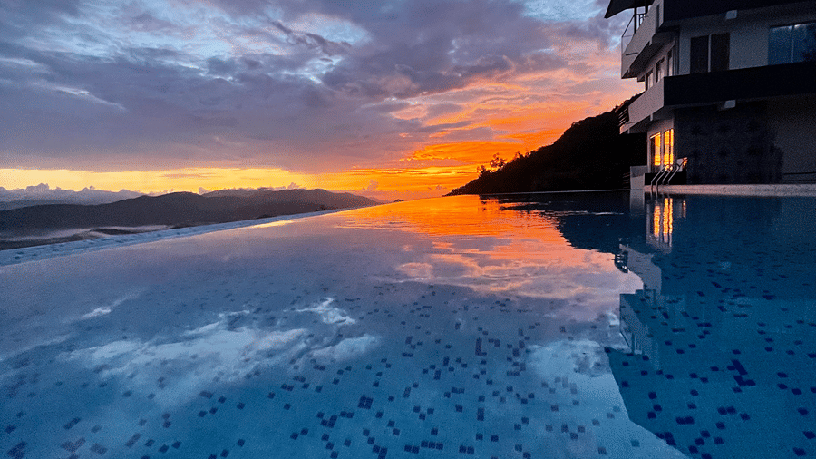 A tiled infinity swimming pool reflecting a colorful sunset sky next to a building | Kollanoor Guest House, Coorg