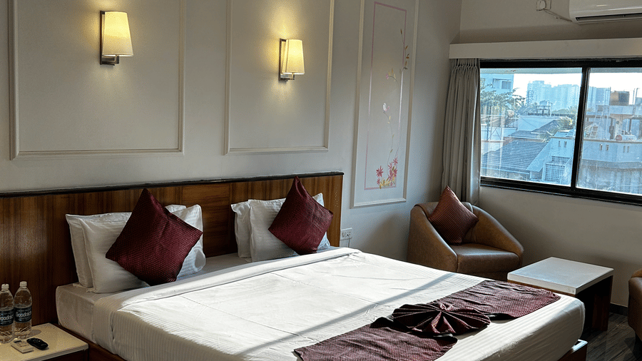  A comfortable guest room at Pravasa Hotel, Kolhapur, featuring a double bed, subtle wall panel moulding, and a wicker armchair.