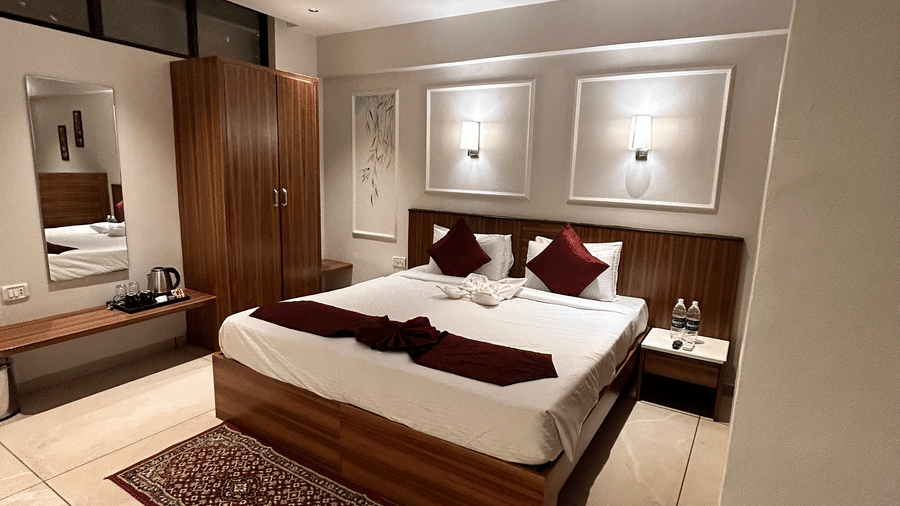 A warm, well-lit standard bedroom featuring a double bed, wooden furniture, and a ceiling fan at Pravasa Hotel, Kolhapur.