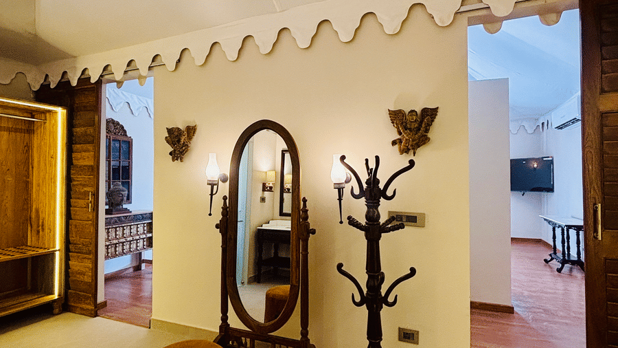 Vana Junior Suite at Surpura Bagh featuring full size vanity mirror and a stool chair in front of it and a wooden piece which holds accessories and a wardrobe with light fixtures. 