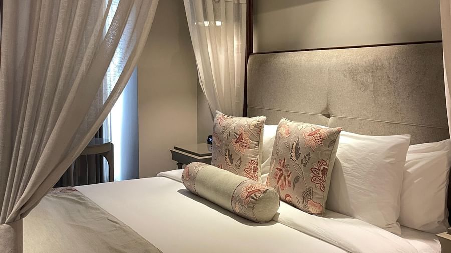 A side view of the four-poster bed in a Premium King Room at The Evren, Vagator. The bed is dressed with a bed runner and decorative pillows, and the white curtains are tied back, letting in natural light.