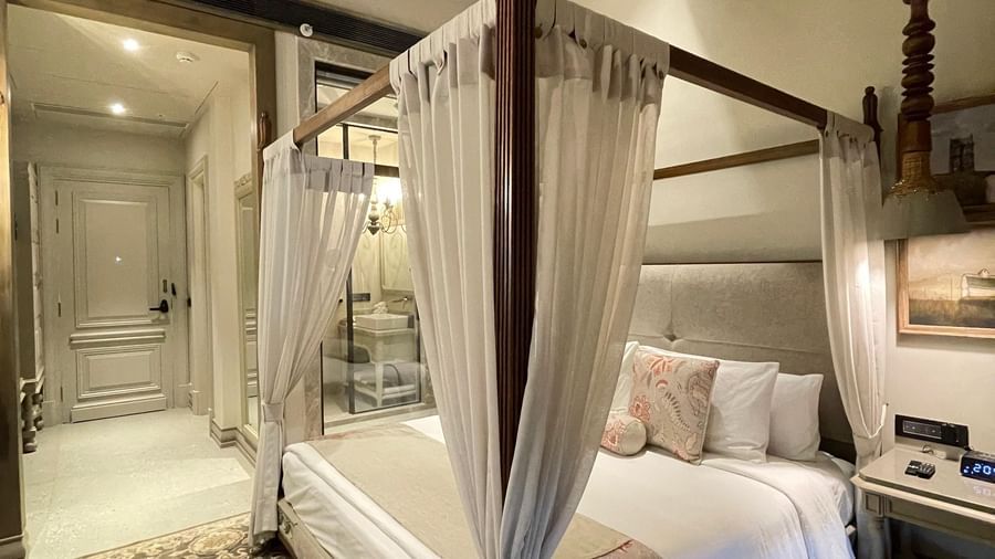An elegant Premium King Room at The Evren, Vagator, featuring a four-poster bed with white curtains, a sloped wooden ceiling with a fan, and a view into the entryway. A patterned rug adds a touch of warmth to the space.