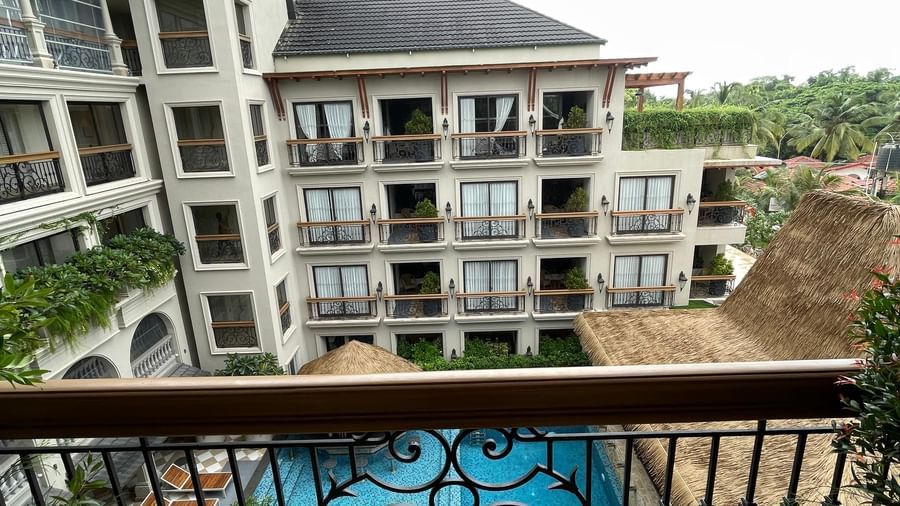 A charming balcony at The Evren, Vagator, with two wicker chairs and a small table, offering a beautiful overhead view of the hotel's swimming pool and the surrounding buildings.