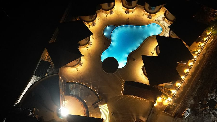 An aerial night view of The Qualia Resort Club & Brewery, Udaipur capturing a pool area, illuminated with warm lights and surrounded by lush greenery.