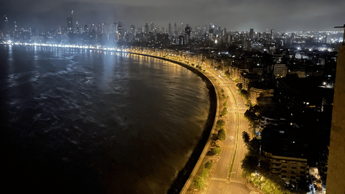 An aerial night view of a coastal road with streetlights along the shoreline and city buildings nearby.