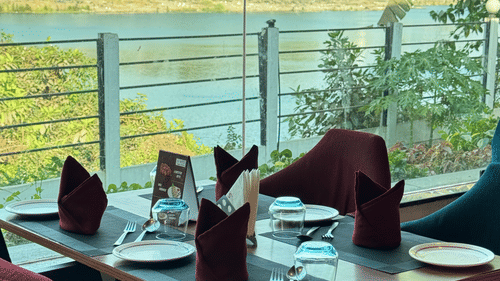 Four seater dining space glass-walled near by a lake surrounded with trees at VITS Kamats Resort, Silvassa