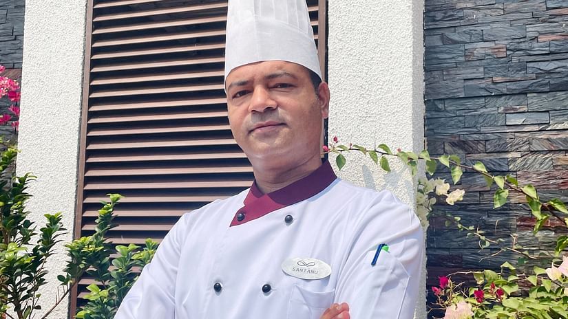 A smiling chef in a white uniform and toque standing outdoors in front of a modern building.