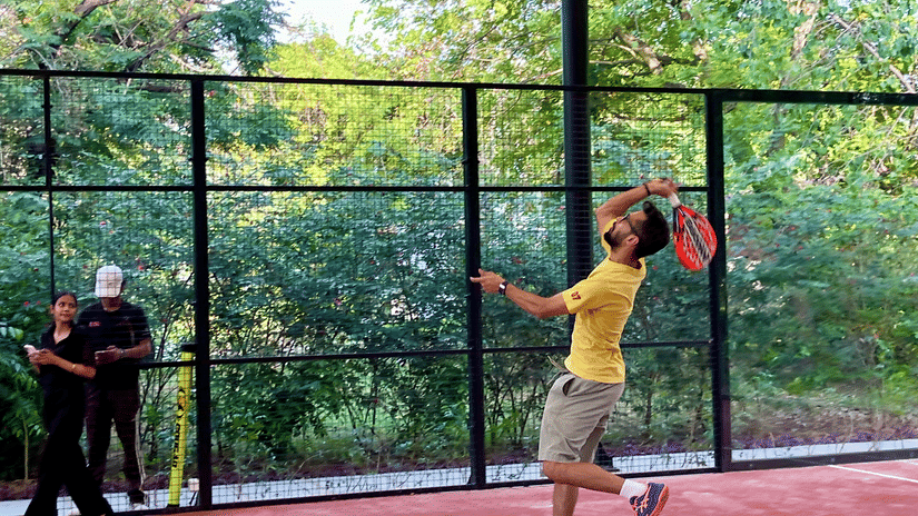 A man in a yellow shirt jumping to hit a ball on a red court surrounded by greenery during a session of padel in Gurgaon at Karma Lakelands