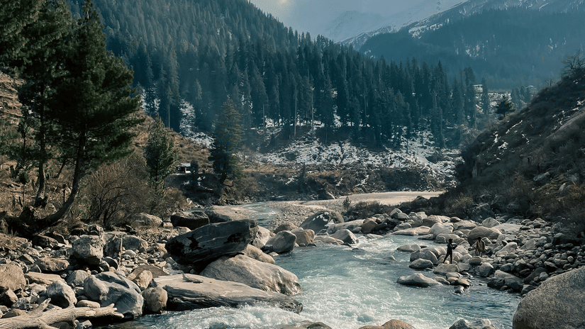A river flowing through rocks and boulders with pine trees and snow-capped mountains seen in the distance.