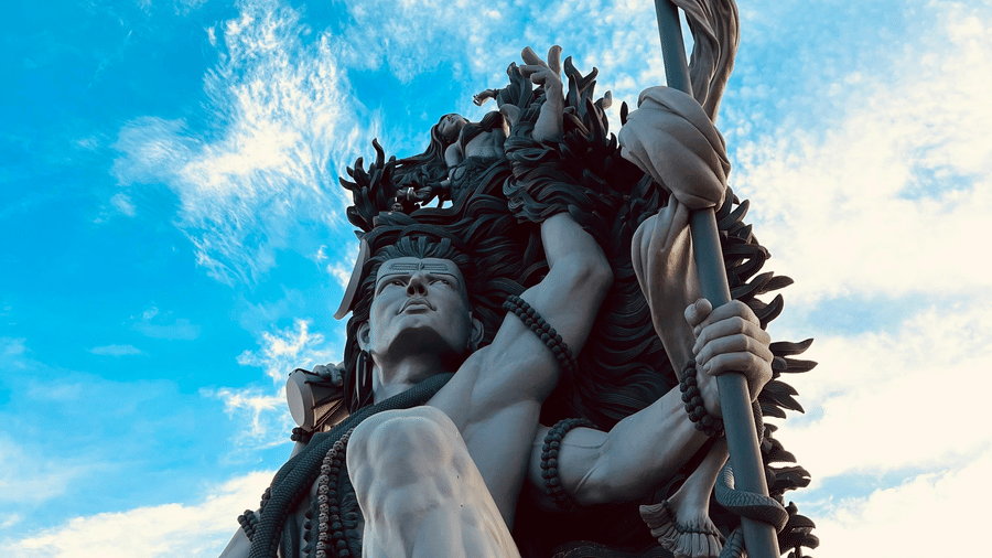 A tall statue of Lord Shiva and a trident, viewed from below against a bright blue sky with scattered clouds.